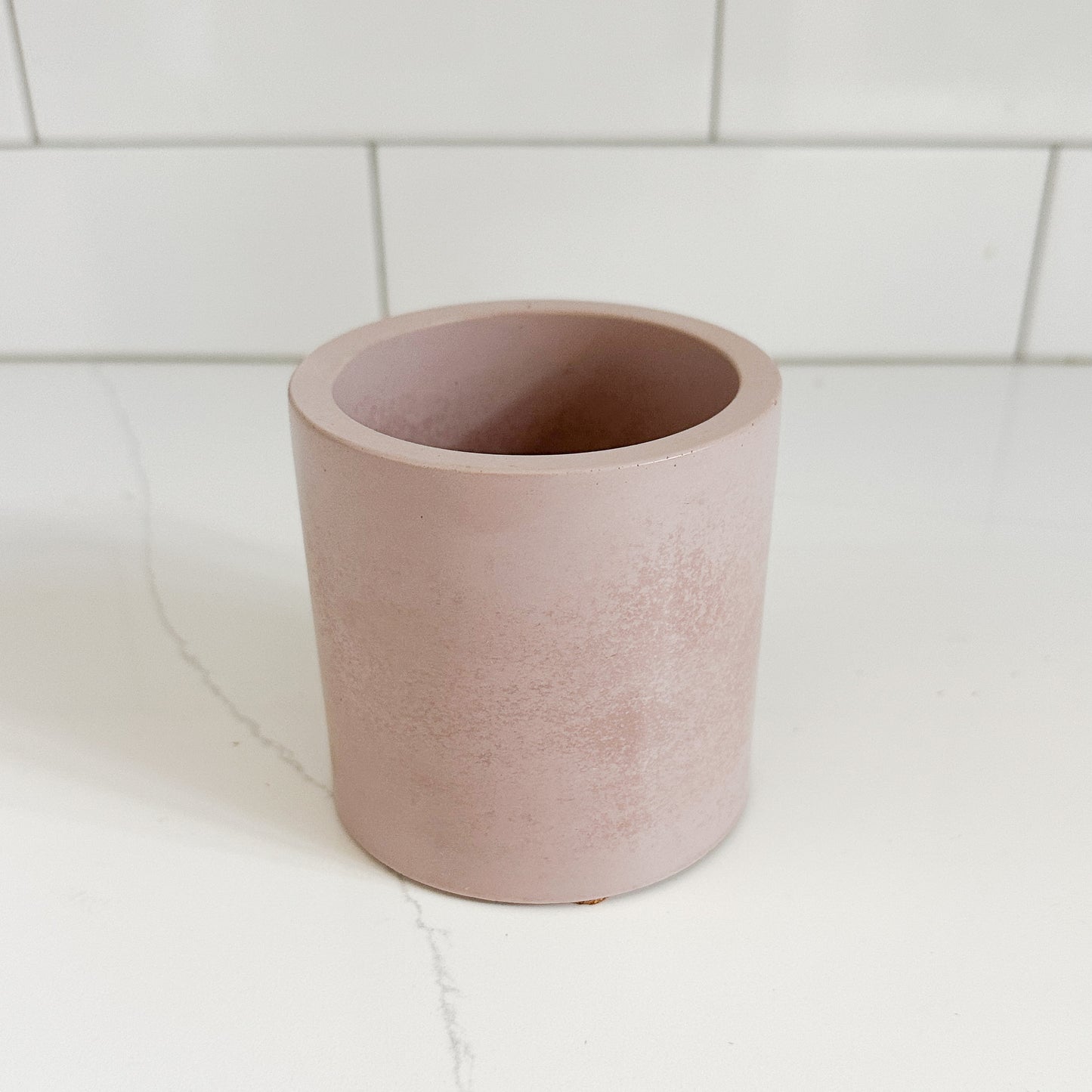A minimalist, open-top cylindrical planter in a dusty rose pink color, set on a white marble countertop with a white subway tile backsplash.