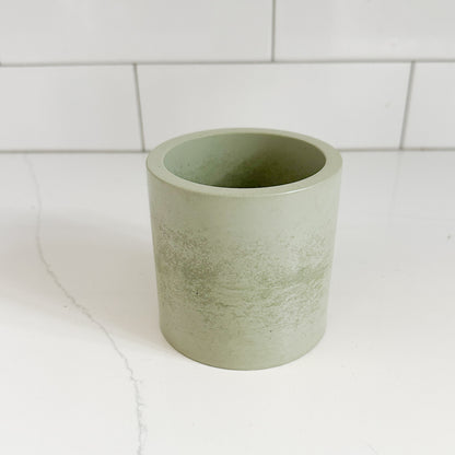 A light sage green cylindrical cup or small planter with a textured, matte finish, set on a white marble surface with a white subway tile background.