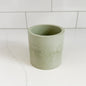 A light sage green cylindrical cup or small planter with a textured, matte finish, set on a white marble surface with a white subway tile background.