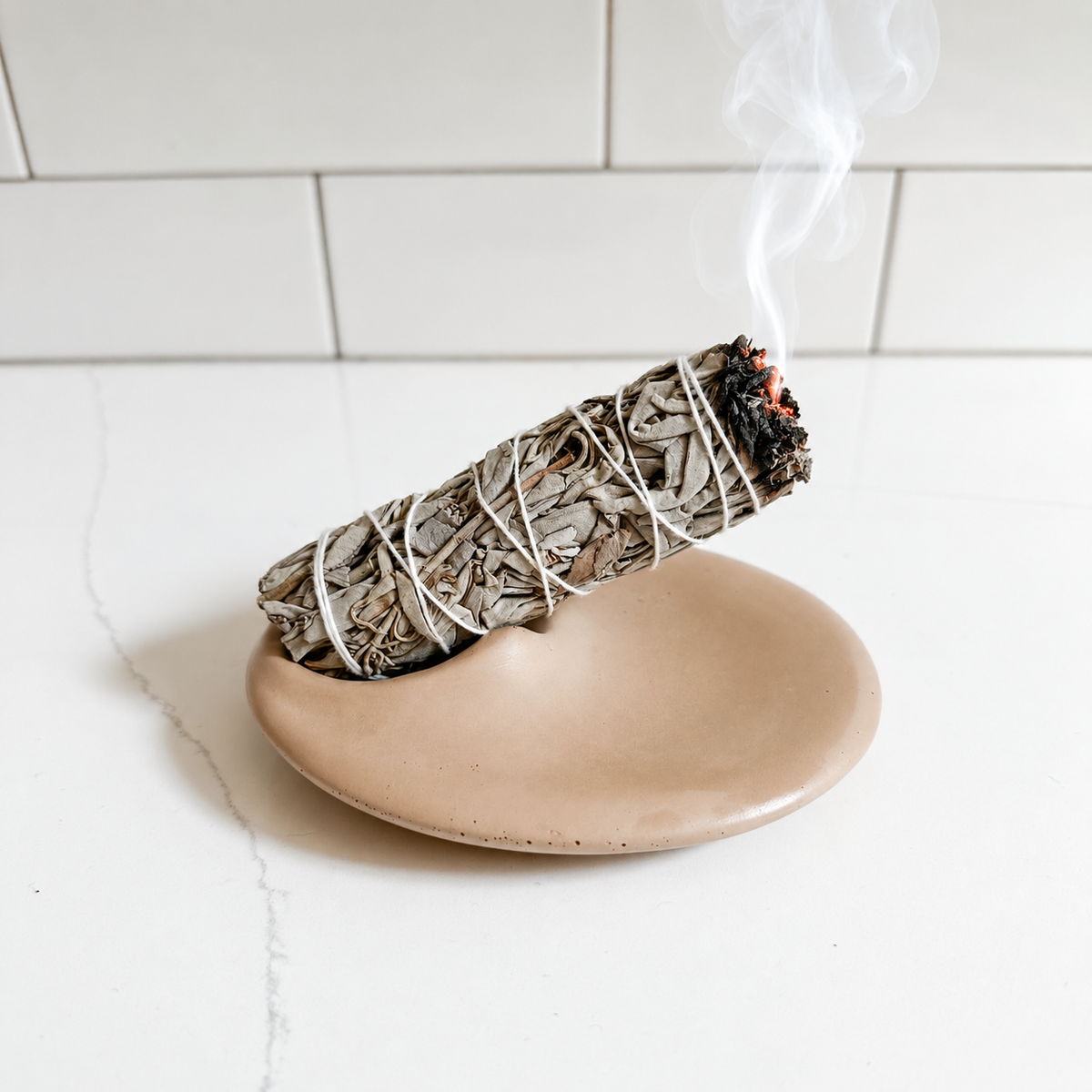 A smudge bundle wrapped in white string, burning with visible smoke, resting on a round concrete incense holder against a white tiled background.